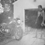 Mężczyzna przy zabytkowym motocyklu przed garażem, trzyma ściereczkę